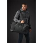 Person wearing a gray jacket holds a large black duffel bag against a dark background.