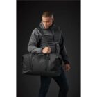 Person in gray jacket and jeans holding a large black duffel bag.