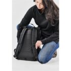 Person crouching, adjusting a black backpack, wearing a black sweater and blue jeans.