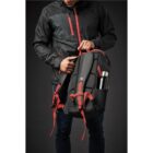 Person holding a backpack with red straps, wearing a black jacket.