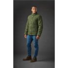 Person in green winter jacket and jeans standing against a dark background.
