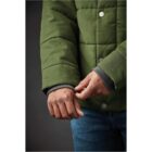Person adjusting sleeve of green padded jacket, wearing blue jeans against dark background.