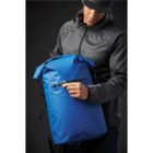 Person in a dark jacket holding a blue backpack against a dark background.
