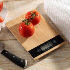 Two tomatoes on a digital kitchen scale next to a knife and towel on a wooden surface.