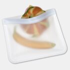Reusable silicone bag containing a sandwich with lettuce and a banana, shown against a plain background. The bag is partially transparent, revealing the food items inside.
