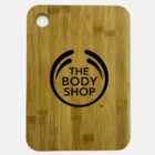 A rectangular bamboo cutting board with rounded edges. It features a circular hole in the top left corner and is printed with The Body Shop logo in black in the center. The background is a plain white surface.