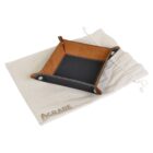 A small black and brown leather tray with visible stitching, placed on a light-colored drawstring cloth bag with AGRADE printed on it. The tray has raised sides and secured corners forming a rectangular shape.