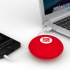 A red, circular power bank with the Uniqlo logo charges a smartphone connected to a MacBook via USB cables. The devices are placed on a white surface.