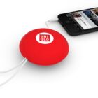 A red circular device with a Uniqlo logo is connected to a black smartphone via a white cable. The smartphone screen displays various apps and media. The setup is on a white surface.