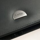 A close-up of a black leather object with a metallic half-circle badge displaying the word Orbit in black font. The surface has a smooth texture with subtle stitching visible along the edge.