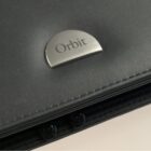 Close-up of a gray leather surface with a metallic semicircular badge displaying the word Orbit. The stitching along the edge is visible, as well as two snap buttons partially seen below.