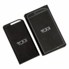 A sleek black portable charger with the Tumi logo is placed next to its matching box. The box features a minimalist design with a white outline of the charger and the Tumi logo.