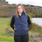 Young woman in a blue vest stands outdoors near a river and rocky hillside, hands in pockets.