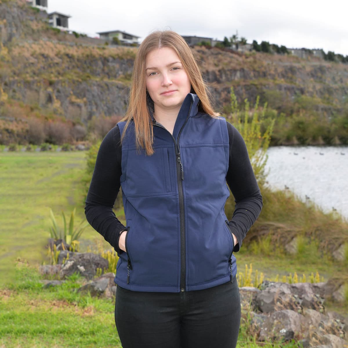Young woman in a blue vest stands outdoors near a river and rocky hillside, hands in pockets.
