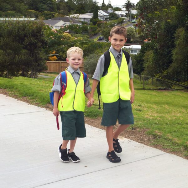 Two young boys walk hand in hand on a sidewalk, wearing bright yellow safety vests and green shorts. One boy has a blue backpack, and the other has a red one. The background shows a neighborhood with trees and houses.
