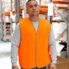 A person wearing a bright orange safety vest and a gray long-sleeve shirt stands in a warehouse. Shelves with boxes are visible in the background.