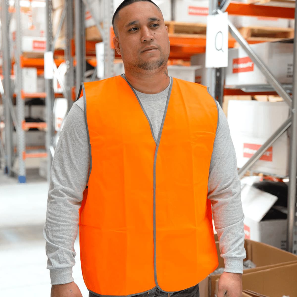 A person wearing a bright orange safety vest and a gray long-sleeve shirt stands in a warehouse. Shelves with boxes are visible in the background.