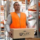 A person wearing an orange safety vest holds a Uflex cardboard box in a warehouse. Shelves with various boxes are visible in the background.