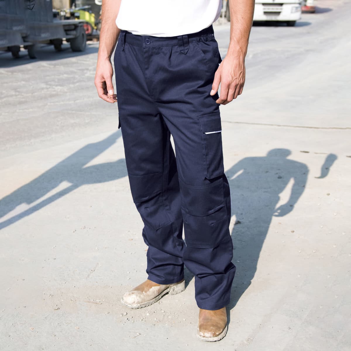A person standing on a paved area wearing navy blue cargo pants and brown work boots. The persons upper body is not visible. The background has a blurry industrial setting with vehicles and equipment. Shadows from the sunlight are visible on the ground.