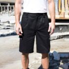 Person wearing black work shorts and a white T-shirt stands outdoors on a construction site. Heavy machinery and concrete debris are visible in the background.