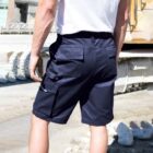 A person wearing dark blue cargo shorts and a white T-shirt standing outdoors, with construction equipment and debris visible in the background. They are facing away from the camera.