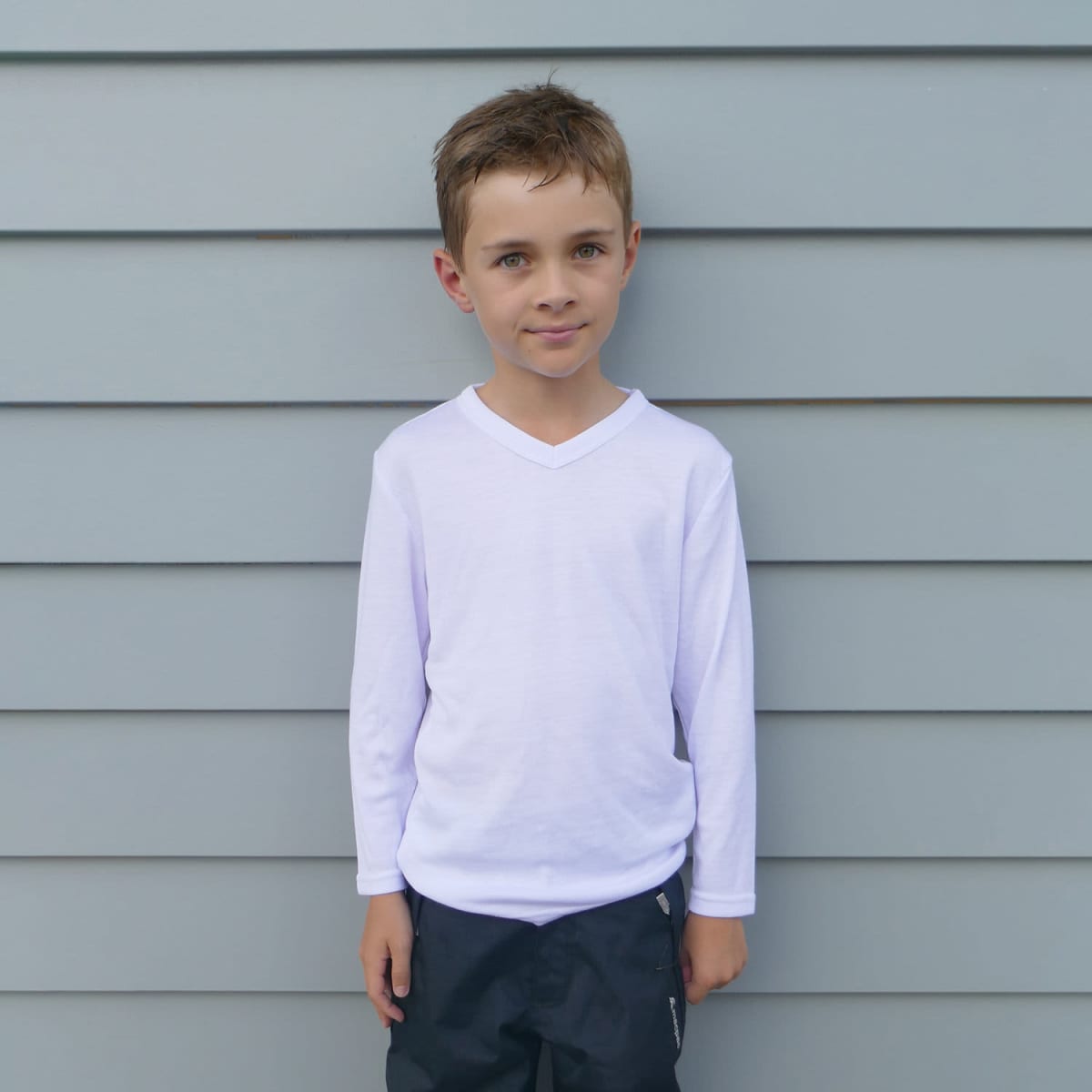 A young boy stands in front of a light gray paneled wall. He is wearing a white long-sleeve shirt and black pants, with his hair neatly styled. He looks directly at the camera.