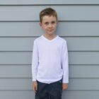 Young boy standing against a gray paneled wall, wearing a white long-sleeve shirt and dark pants, with hands resting at his sides.