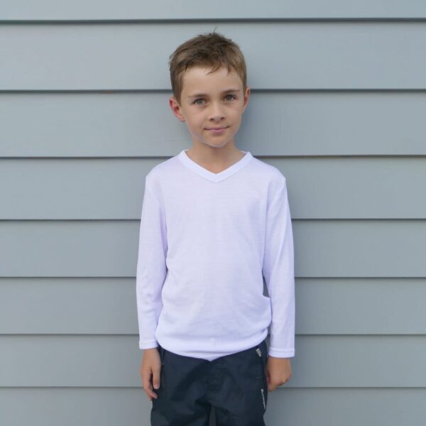 Young boy standing against a gray paneled wall, wearing a white long-sleeve shirt and dark pants, with hands resting at his sides.