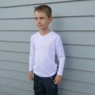 Young boy in a white long-sleeve shirt and dark pants stands in front of a gray, horizontally paneled wall, looking to the side.