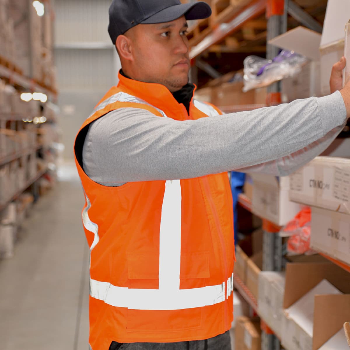 A person in an orange reflective vest and cap is organizing shelves in a warehouse. They are holding a box and appear to be focused on their task. Rows of shelves filled with various boxes and items are visible in the background.