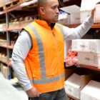 A man wearing an orange safety vest and gray long-sleeve shirt is arranging boxes on a shelf in a warehouse. He stands in an aisle filled with various stacked packages and containers.