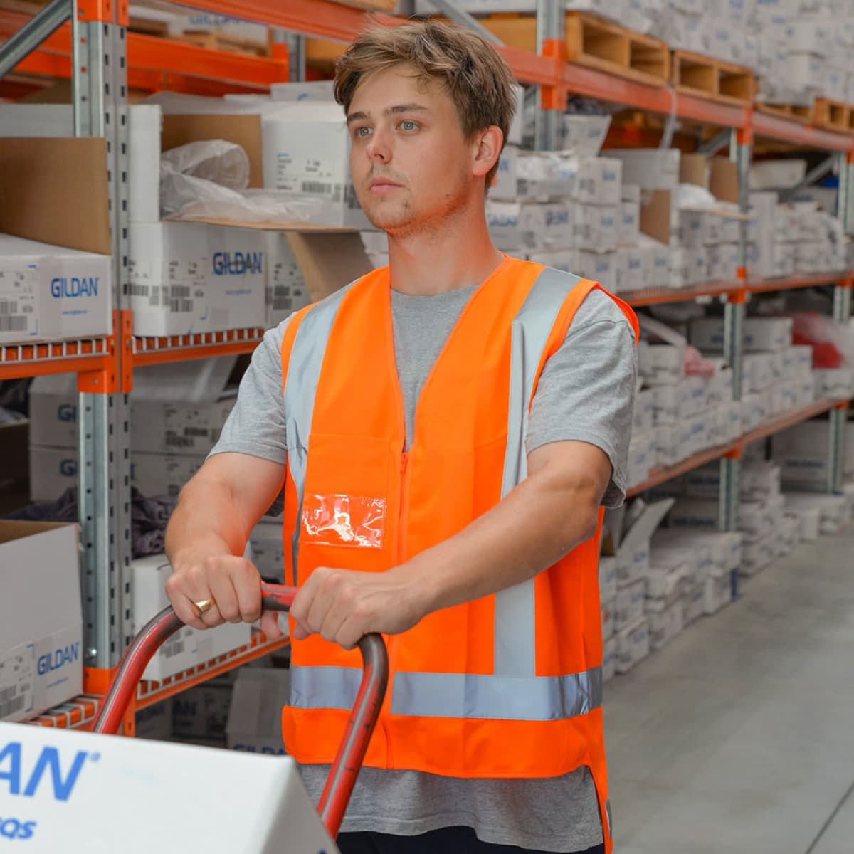A person wearing an orange safety vest pushes a cart down an aisle in a warehouse. The shelves are filled with boxes and various items. The individual has short hair and wears a gray T-shirt under the vest.