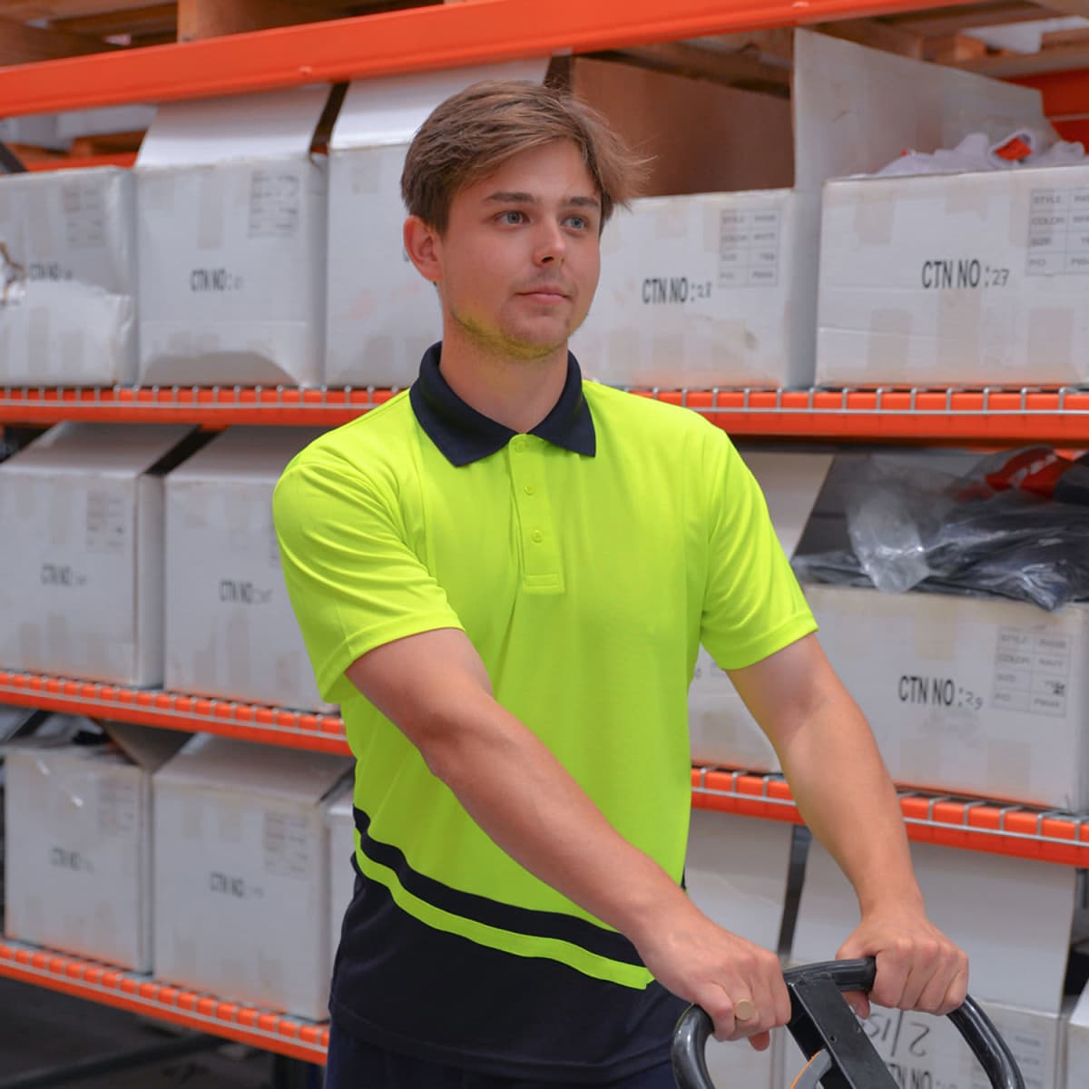 A man in a bright yellow shirt stands in a warehouse with shelves and boxes around. He is holding onto a handle of a cart or trolley, looking focused and prepared for work.