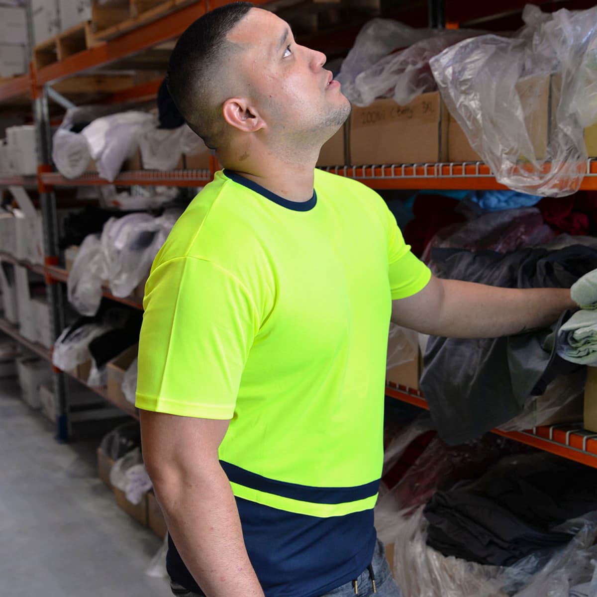 A man in a bright neon yellow shirt stands in a warehouse, looking up. He is near shelves stacked with boxes and plastic-covered items.