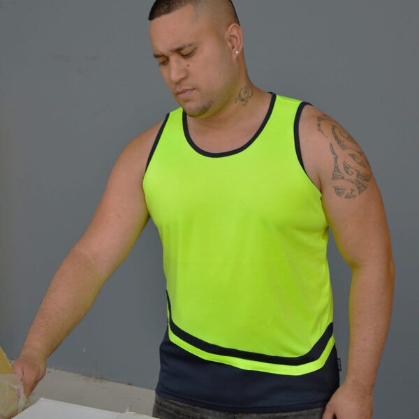 A person with tattoos wearing a bright yellow tank top and dark shorts stands against a gray background, looking down at something off-camera.