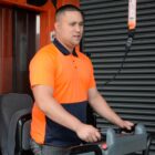 A person wearing an orange and navy polo shirt operates machinery indoors. They have a focused expression, and various control elements are visible in the foreground.