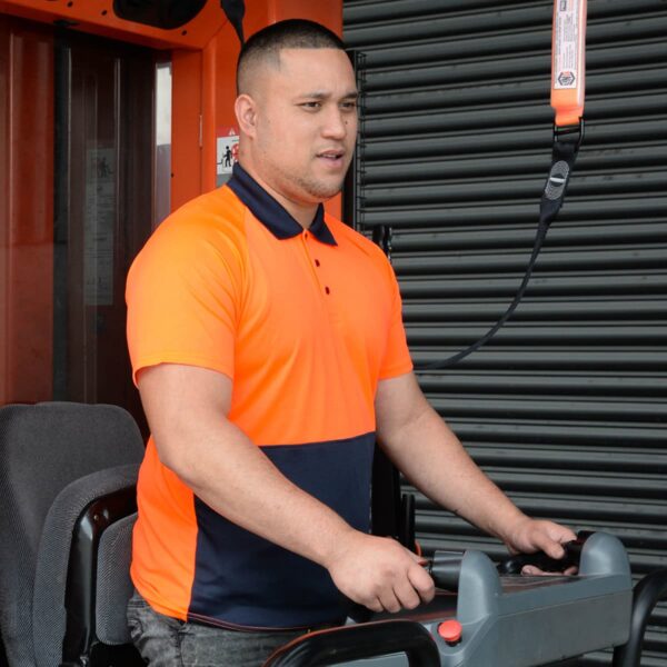 A person in an orange and navy shirt operates machinery inside a building. They have short hair and are standing next to a large wheel, focusing on their task.
