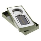 A rectangular keychain with a carbon fiber design and a metallic ring, featuring a gold Lexus logo. It is placed in an open green box with a fitted cushion lining.