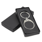 A metal and leather keychain in a black gift box with a lid slightly ajar, showing the contents inside. The keychain has an oval shape with a silver metal ring and a dark leather insert.