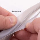 Close-up of hands showing the edges of a white resealable plastic bag. The bags opening mechanism is slightly parted, revealing its closure design. The word Resealable is printed above.