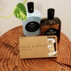 Two bottles of Leif body cleanser, one blue cypress and one lemon myrtle, are placed on a wooden surface. A box labeled Bask & Stow, Byron Bay contains cotton swabs. A green leaf is visible in the background.