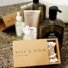 A collection of skincare bottles and containers are displayed on a granite countertop. Visible brands include leif and Bask & Stow. Cotton swabs in an open cardboard box are in the foreground.