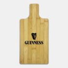 A rectangular wooden cutting board with a handle, featuring the Guinness logo and harp design in black on one side. The board has a smooth, light wood grain finish.