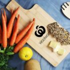 A wooden cutting board with the Woolworths logo. On the board are several fresh carrots with greens, a lemon, slices of seeded bread, and pieces of cheese. A wooden kitchen utensil is visible on the side. The background is a blue surface.