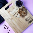 A wooden cutting board with the word Nestle engraved on it holds a small container of dark olives, several whole grain crackers, and seed-covered crackers. Coffee beans are scattered nearby on a pink surface, with a dark cloth partially visible.