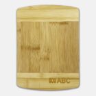 A rectangular bamboo cutting board with lighter wood strips at the top and bottom. The bottom right corner features a logo and the text ABC engraved. The board has a smooth, natural wood grain pattern.
