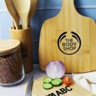 A kitchen scene with wood utensils in a holder, a jar of beans, and cutting boards. One board is branded The Body Shop, and the other shows ABC with sliced cucumber, onion, and tomato on a counter with a knife. Blue tiled background.