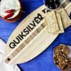A wooden cutting board with the word QUICKSILVER is surrounded by dips, red chili peppers, black olives, and assorted crackers on a blue surface.