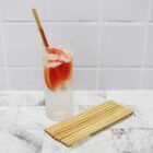A tall glass with a refreshing pink drink, ice cubes, and a bamboo straw is on a marble surface. Next to it lies a set of bamboo straws and a cleaning brush. The background features white tiled walls.