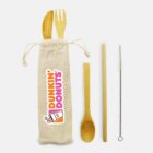 A reusable bamboo cutlery set featuring a fork, knife, spoon, and chopsticks alongside a straw with a cleaning brush. The items are placed next to a fabric pouch labeled with the Dunkin Donuts logo.